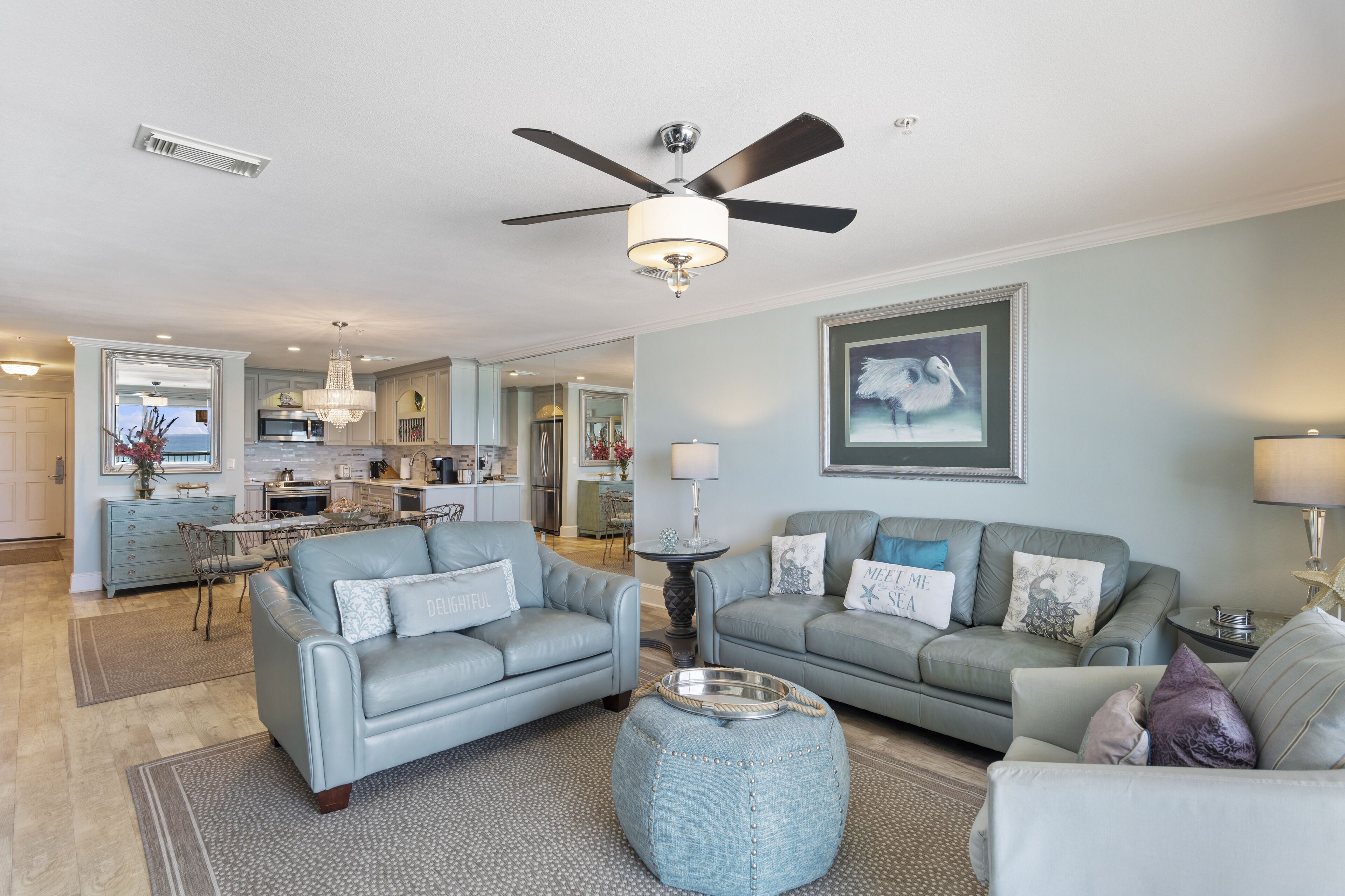 A spacious living room with two sofas, a coffee table, and a ceiling fan. A kitchen is visible in the background with a dining area.