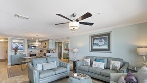 A spacious living room with two sofas, a coffee table, and a ceiling fan. A kitchen is visible in the background with a dining area. - Newly Renovated Condo 305, Your Luxurious BEACH FRONT Paradise - Crystal Dune (Destin)