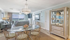 A spacious living room with a glass dining table surrounded by wicker chairs, a large chandelier, and a decorative cabinet with glass doors. - Newly Renovated Condo 305, Your Luxurious BEACH FRONT Paradise - Crystal Dune (Destin)