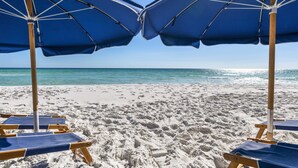 Deux parasols de plage bleus et des chaises longues en bois sur une plage de sable, avec une eau bleu clair en arrière-plan.