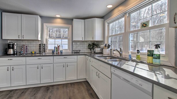 A modern kitchen with white cabinets, a large window with a view of trees, and a dishwasher. - Hampton 'Quiet Corner Cottage' w/ Fireplace & Yard (Hampton)