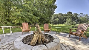 A stone fire pit surrounded by Adirondack chairs, set on a stone patio with a grassy lawn and trees in the background. - Hampton 'Quiet Corner Cottage' w/ Fireplace & Yard (Hampton)