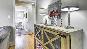 A modern living room with a grey sofa, a wooden cabinet with a mesh door, and a dining area with a white table and chairs. - Hampton 'Quiet Corner Cottage' w/ Fireplace & Yard (Hampton)