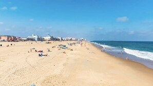 A wide sandy beach with umbrellas and people, a clear blue sky, and buildings in the background.