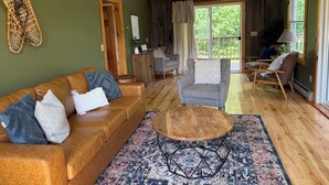 A cozy living room with a brown leather sofa, a grey armchair, and a wooden coffee table. There is a patterned rug on the floor and a large window with curtains.