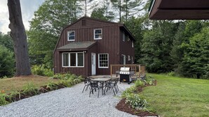 A two-story wooden cabin with a deck, a barbecue grill, and a gravel driveway.