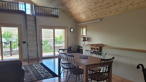 Une pièce spacieuse avec un plafond en bois, une table à manger avec des chaises, un canapé, une échelle et un balcon donnant sur un paysage verdoyant.