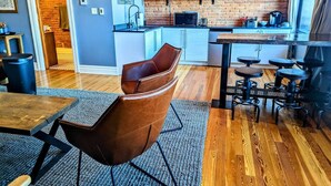 A modern kitchen with a brick wall, wooden flooring, and a dining area with a leather chair and stools. - Swanky Downtown Loft (Waverly)