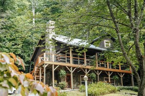 Una cabaña rústica con terraza de madera y amplias ventanas, rodeada de un bosque denso.
