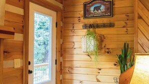 Interno di una baita con una porta di legno, un cartello di benvenuto, una pianta sospesa, una lampada e un tappeto con decorazioni di animali.