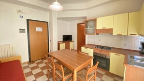 A compact kitchen with a dining table, chairs, a stove, and yellow cabinets. - Appartamento Adriana 3 - Carraro Immobiliare Jesolo (Lido di Jesolo)