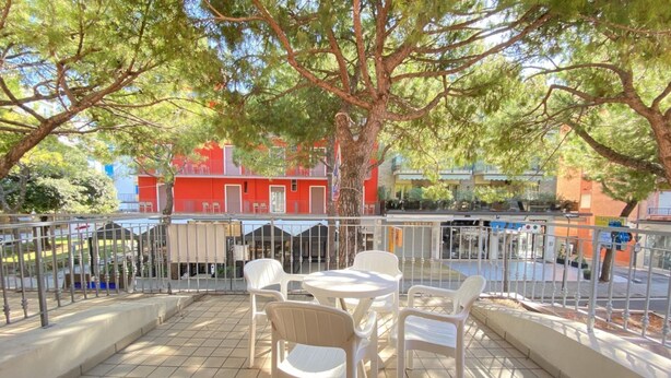 An outdoor patio with white plastic chairs and tables, a metal railing, and a view of a red building in the background. - Appartamento Adriana 3 - Carraro Immobiliare Jesolo (Lido di Jesolo)
