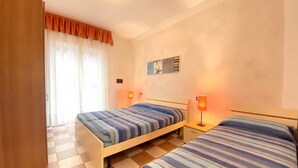 A hotel room with two beds, a checkered floor, a window with curtains, and a wall clock. - Appartamento Adriana 3 - Carraro Immobiliare Jesolo (Lido di Jesolo)