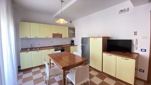 Una cocina compacta con una mesa de madera, sillas blancas, un televisor y armarios de tonos claros.