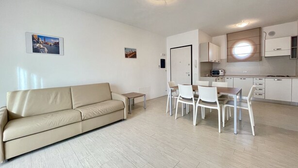 A modern living room with a beige sofa, a dining table with chairs, and a kitchen area with cabinets and a microwave. - Appartamento Treporti 4 Cavallino- Carraro Immobiliare Jesolo (Cavallino)