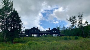 Une grande maison aux couleurs sombres, dotée de nombreuses cheminées, entourée d’une végétation luxuriante et d’un champ de fleurs sauvages.