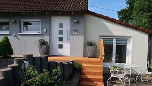 Ein modernes Haus mit Ziegeldach, einer weißen Tür und einem Balkon mit Holzboden und Metallstühlen.