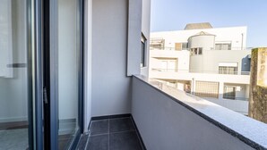 A narrow balcony with a glass door, a tiled floor, and a view of a modern building with multiple balconies and windows.