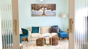 Una sala de estar con un sofá color beige, un sillón azul verdoso y un cuadro de un árbol. Hay dos mesitas de mimbre y una lámpara.