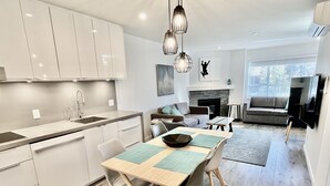 Eine moderne Küche mit weißen Schränken, ein Essbereich mit einem Holztisch und Stühlen, ein Kamin und ein Wohnzimmer mit Sofa und Fernseher.
