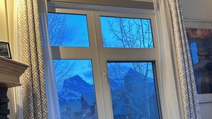A living room with a beige sofa, patterned curtains, and a view of a snowy mountain landscape through a large window. - SUNNY BRIGHT King Bed Suite Full Kitchen Free Parking Balcony BBQ Gym (Canmore)