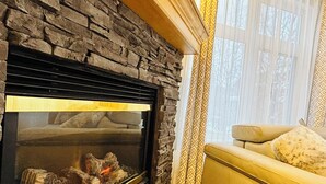 A stone fireplace with a glass door, a sofa with a fluffy throw, and patterned curtains. - SUNNY BRIGHT King Bed Suite Full Kitchen Free Parking Balcony BBQ Gym (Canmore)