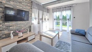 Ein modernes Wohnzimmer mit einem Flachbildfernseher an einer Ziegelwand, einem Holztisch, einem grauen Sofa und einem großen Fenster mit durchsichtigen Vorhängen.