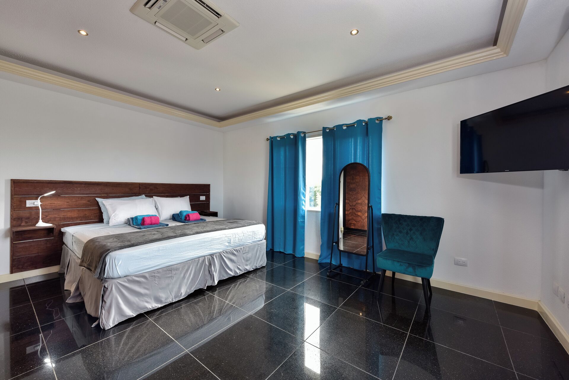 A hotel room with a bed, a nightstand, a desk lamp, a chair, a TV, and a window with blue curtains.