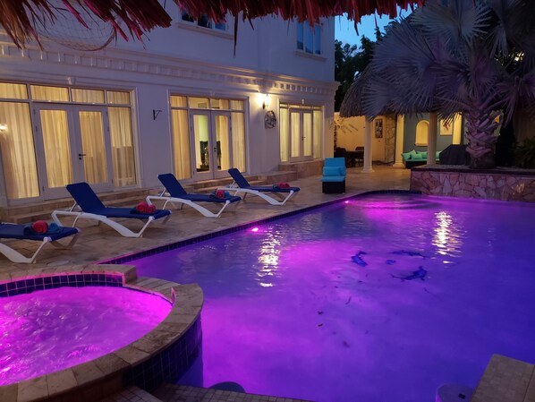 A swimming pool with lounge chairs and a hot tub, surrounded by a stone wall and palm trees.