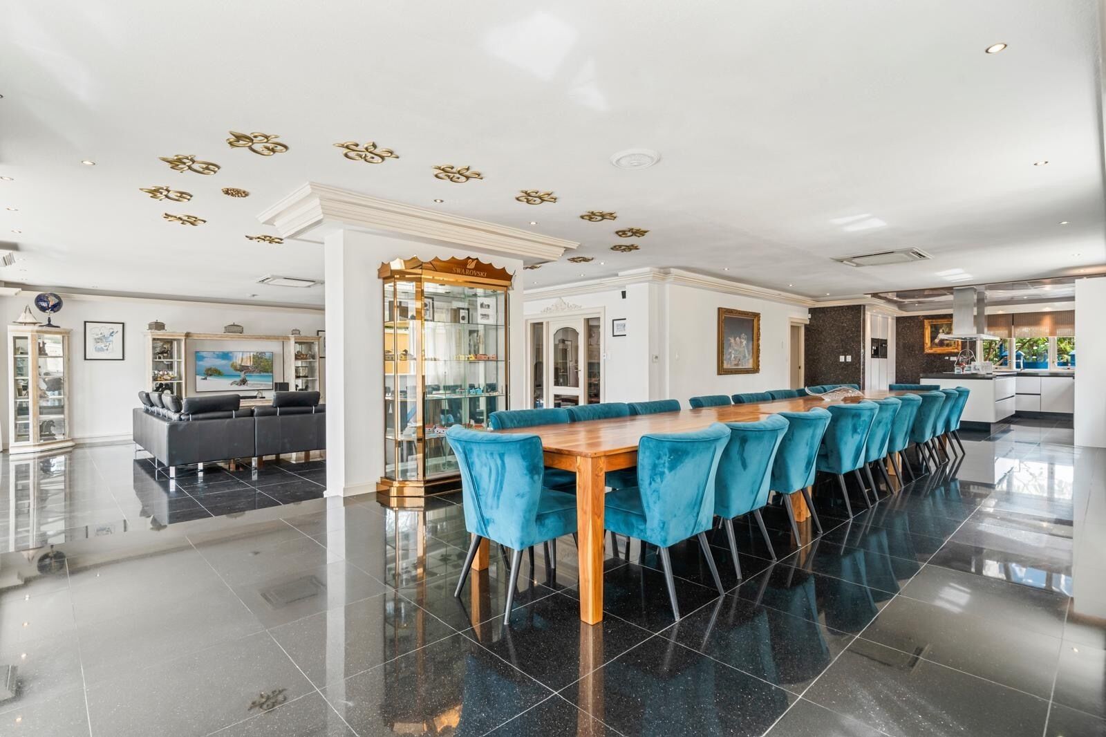 Uma sala de jantar espaçosa com uma mesa de madeira comprida e cadeiras estofadas em azul, uma cozinha moderna e uma sala de estar com sofá e sistema de entretenimento.