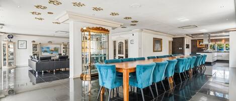 Uma sala de jantar espaçosa com uma mesa de madeira comprida e cadeiras estofadas em azul, uma cozinha moderna e uma sala de estar com sofá e sistema de entretenimento.