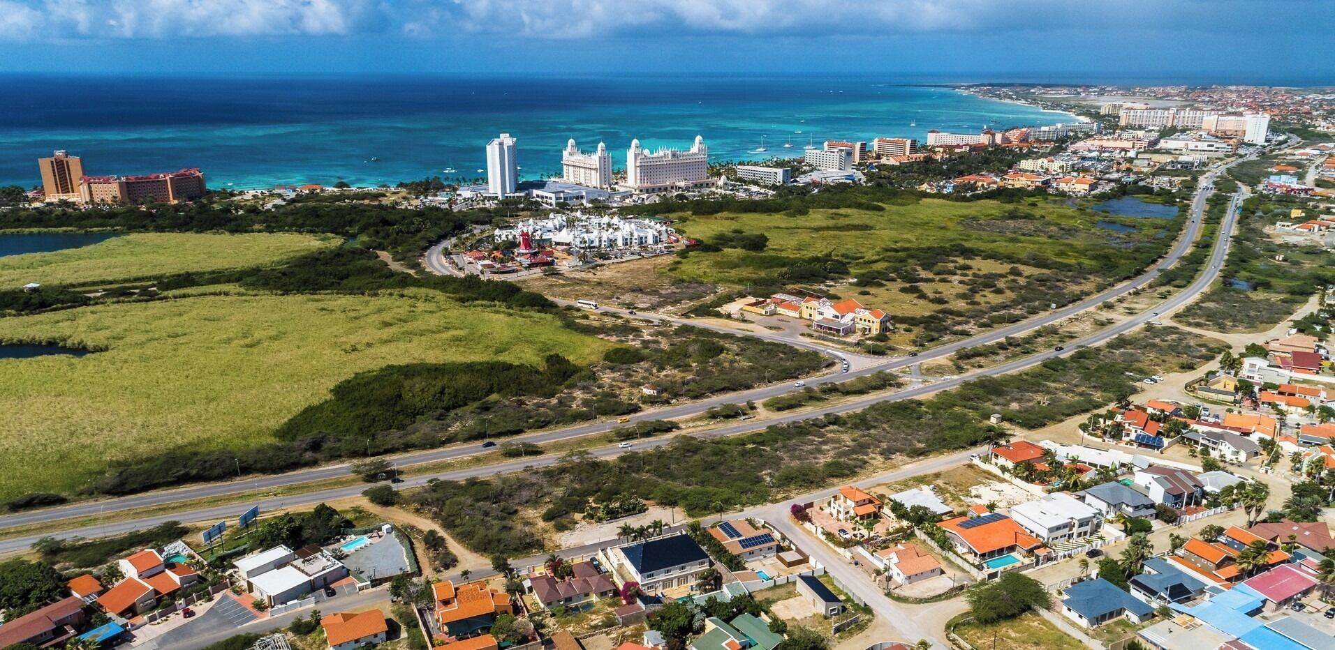 An aerial view of a coastal town with a mix of residential and commercial buildings, a prominent hotel with a white facade, and a clear view of the ocean.