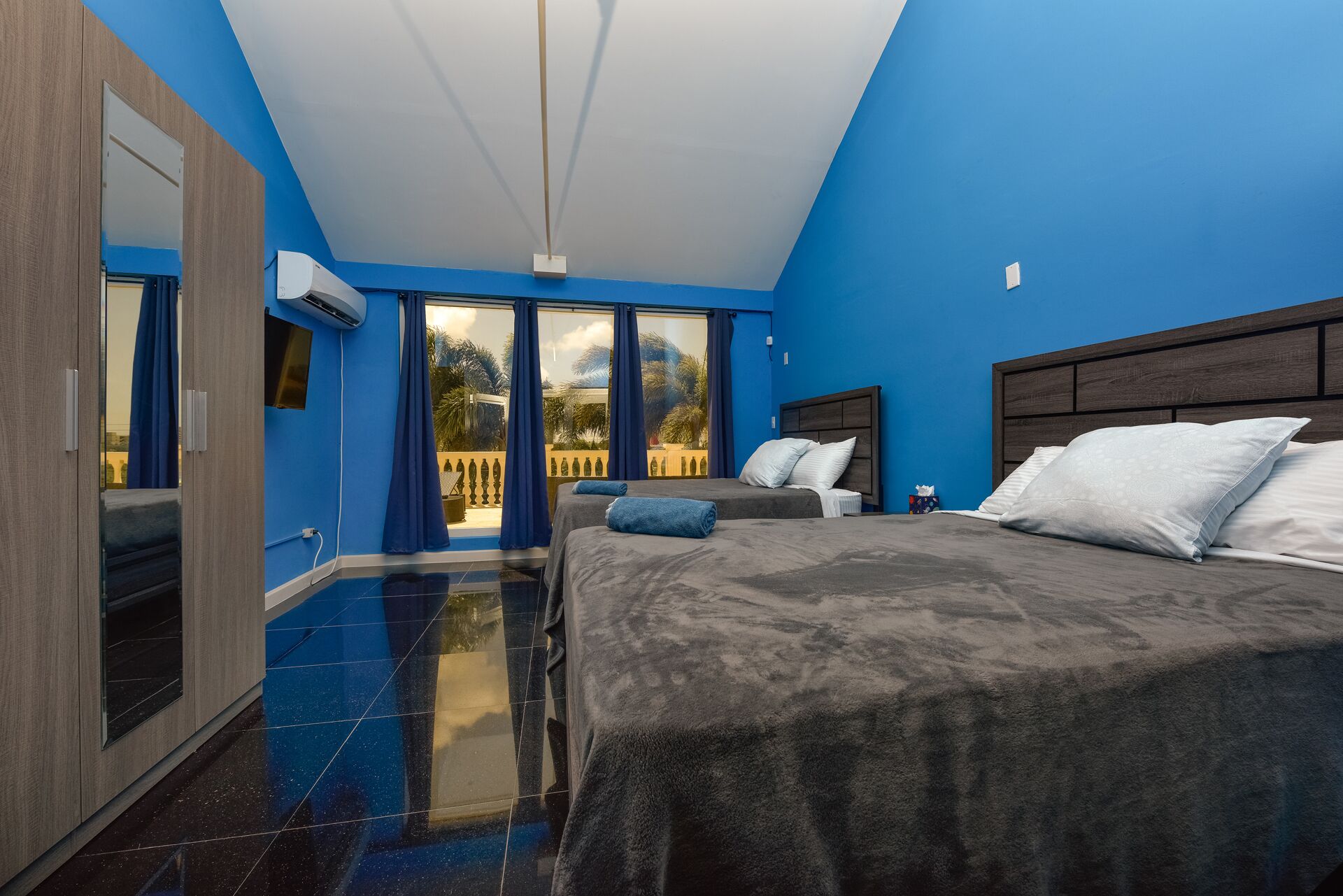 A modern bedroom with a large bed, a balcony with blue curtains, and a view of palm trees.