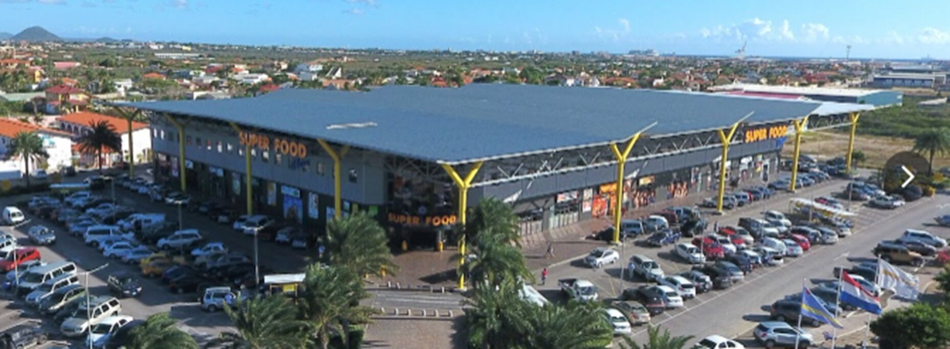 Un edificio grande con techo plano y múltiples accesos, rodeado por un estacionamiento y palmeras.