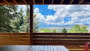 Una terraza de madera con una mesa y sillas, con vistas a un paisaje montañoso con árboles y cielo despejado.