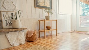 Una habitación con piso de madera, una cesta tejida, una silla de madera, un tapiz tejido y una planta en una maceta estampada.