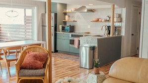 Una sala moderna con un sofá, una silla de mimbre, una mesa redonda con flores y una zona de cocina con refrigerador, horno y microondas.