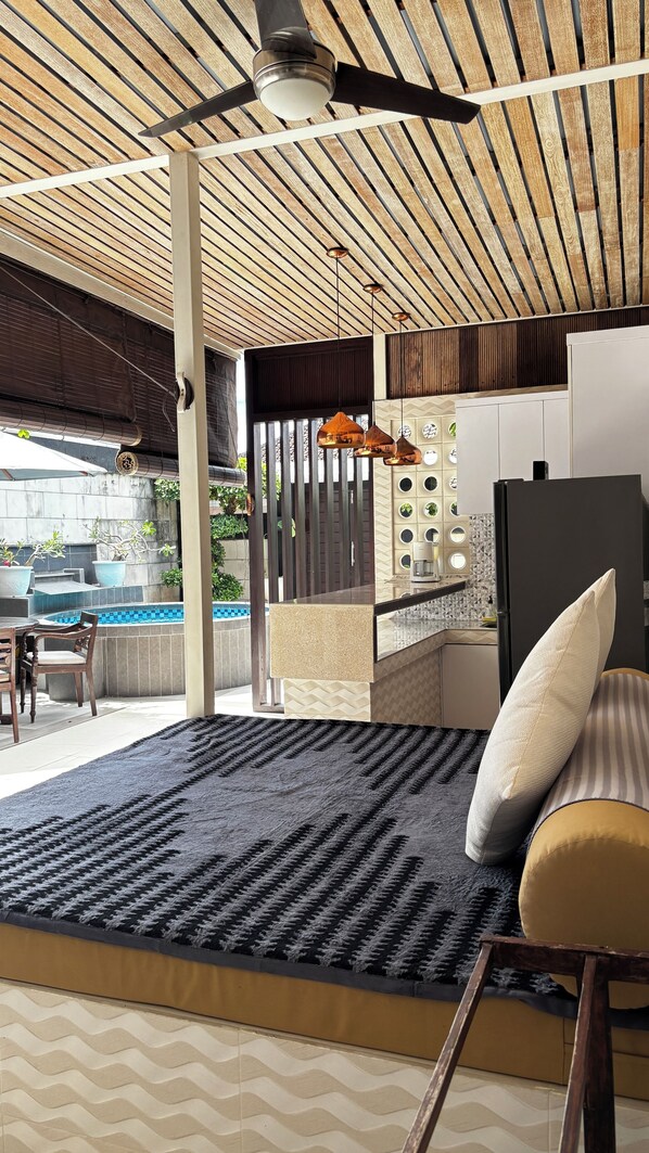 Una sala de estar moderna con un ventanal grande, ventilador de techo y zona de cocina con refrigerador y pileta.
