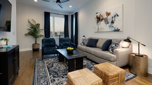 A modern living room with a sofa, chairs, a coffee table, and a floor lamp. There is a large window with curtains, a ceiling fan, and a painting on the wall. - SKYLINE ROOFTOP DECK 4 BEDROOMS KING BED LARGE KITCHEN L2 EV CHARGER PETS GARAGE (Nashville)