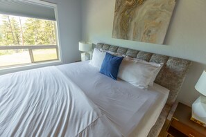 Una cama bien hecha con sábanas blancas y una almohada azul, una obra de arte de mármol en la pared y una ventana con persianas.