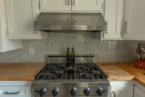 Una cocina con armarios blancos, una campana extractora de acero inoxidable, una estufa a gas con cuatro quemadores y dos botellas de aceite de oliva sobre la encimera.