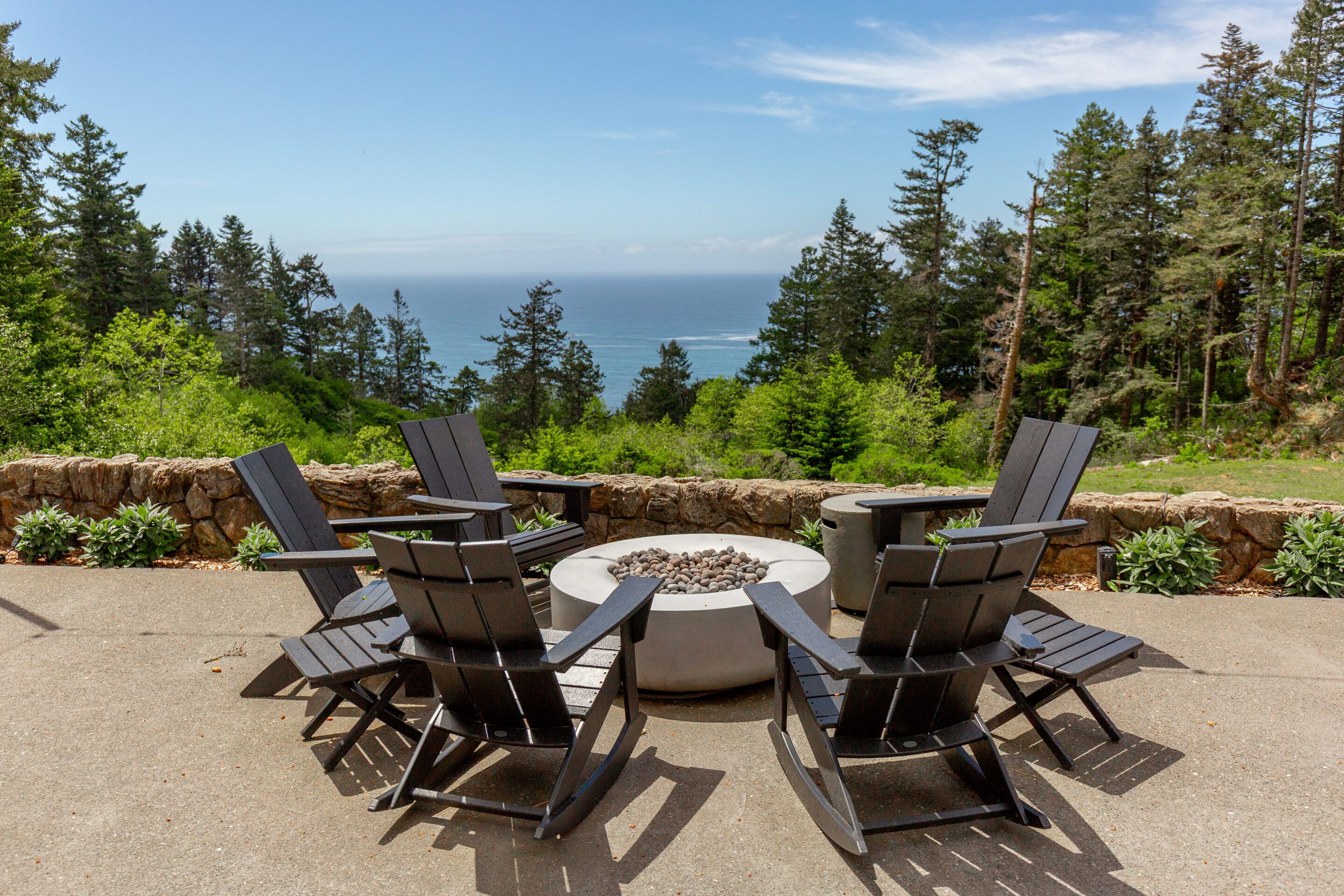 An outdoor seating area with Adirondack chairs and a circular fire pit, overlooking a forested landscape and the ocean.