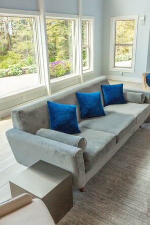 A modern living room with a grey sectional sofa and blue cushions, large windows offering a view of greenery, and a light-colored carpet.