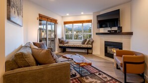 A cozy living room with a sofa, a coffee table, a fireplace, and a large window with a mountain view. - Heart of the Village. Newly renovated , modern décor throughout, Central AC just installed 2024 (Whistler)