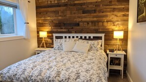 Ein Schlafzimmer mit einer Holzwand, einem Bett mit floralem Bettzeug, zwei Nachttischen mit Lampen und einem Fenster mit Vorhängen.