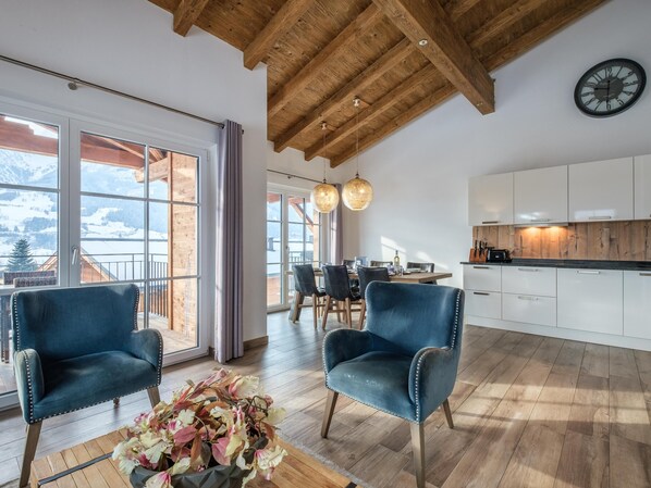 A spacious living area with a wooden ceiling, large windows, a dining area, and a kitchen with white cabinets. - Stylish penthouse with shared sauna and lift (Walchen)