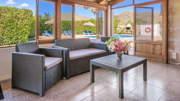 A covered outdoor seating area with wicker furniture, a table, and a view of a pool and mountains. - Vacation Villa 'Can Serra' with Private Pool, Mountain Views, Terrace & Garden (Puerto Pollensa)