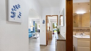 Um corredor com porta de madeira dando para um banheiro, um sofá azul na sala de estar e um quadro na parede.