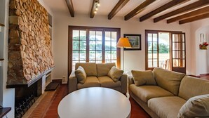 A living room with a stone wall, wooden beams, a sofa, a table, and large windows. - Wonderful Villa “Villa Las Palmeras” with Sea View, Pool, Wi-Fi, A/C, Garden & Terrace (Binibequer)