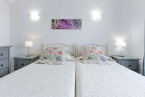 Una cama doble con almohadones florales, dos mesitas de noche con lámparas, una lámpara de pared y un cuadro enmarcado sobre la cama.